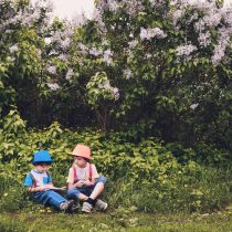 Fotografia raffigurante due bambini seduti in un prato, vestiti con abiti da giardino